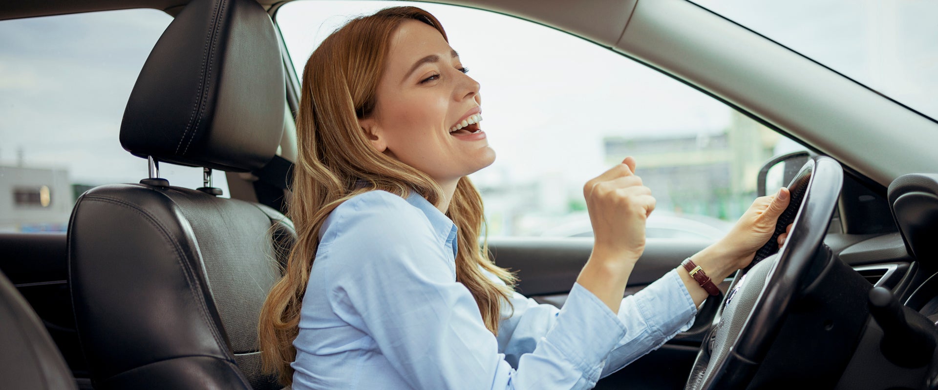 A woman in a car, smiling and singing with enjoyment. She's wearing a blue shirt, hands raised near the steering wheel, exuding happiness and energy.